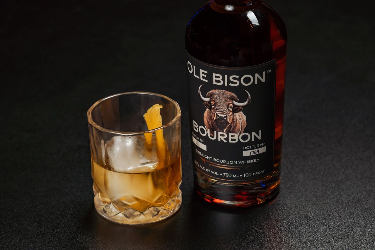 A bottle of Ole Bison Bourbon alongside a glass of whiskey with ice and orange peel.